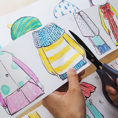 Rankos juodomis žirklėmis kerpa geltonu megztiniu, mėlyna kepure ir šaliku nuspalvintą Little Designer mergaitės figūrą