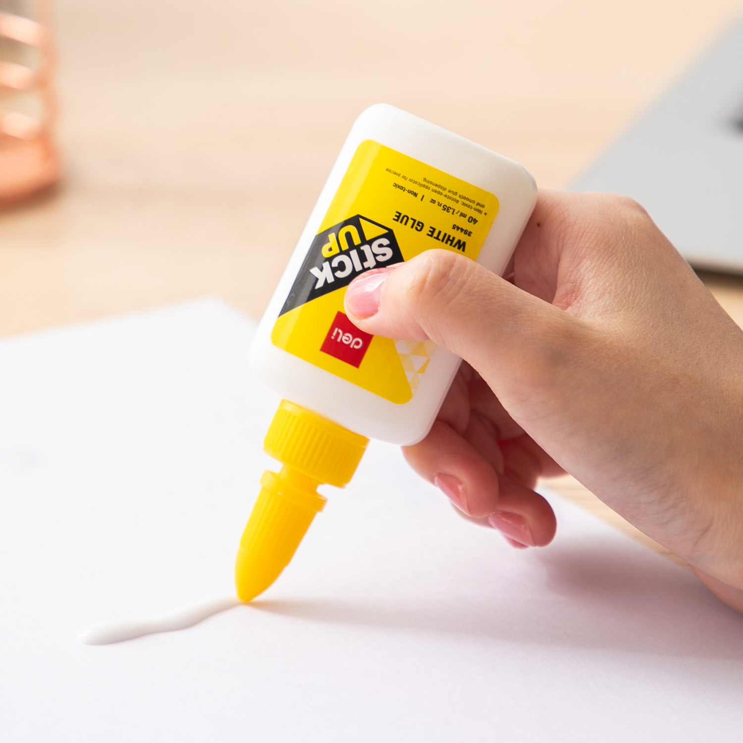 Ranka spaudžia apverstą Deli Stick Up 40 ml buteliuką, o geltonu aplikatoriumi ant balto popieriaus liejama klijų juosta