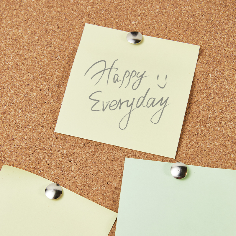 Trys šviesūs lapeliai sidabriniais smeigtukais pritvirtinti prie kamštinės lentos, viršutiniame parašyta Happy Everyday