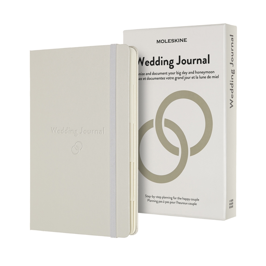 Kreminė Moleskine Wedding Journal knygelė su elastine juosta stovi prieš baltą dėžutę su dviejų žiedų simboliu
