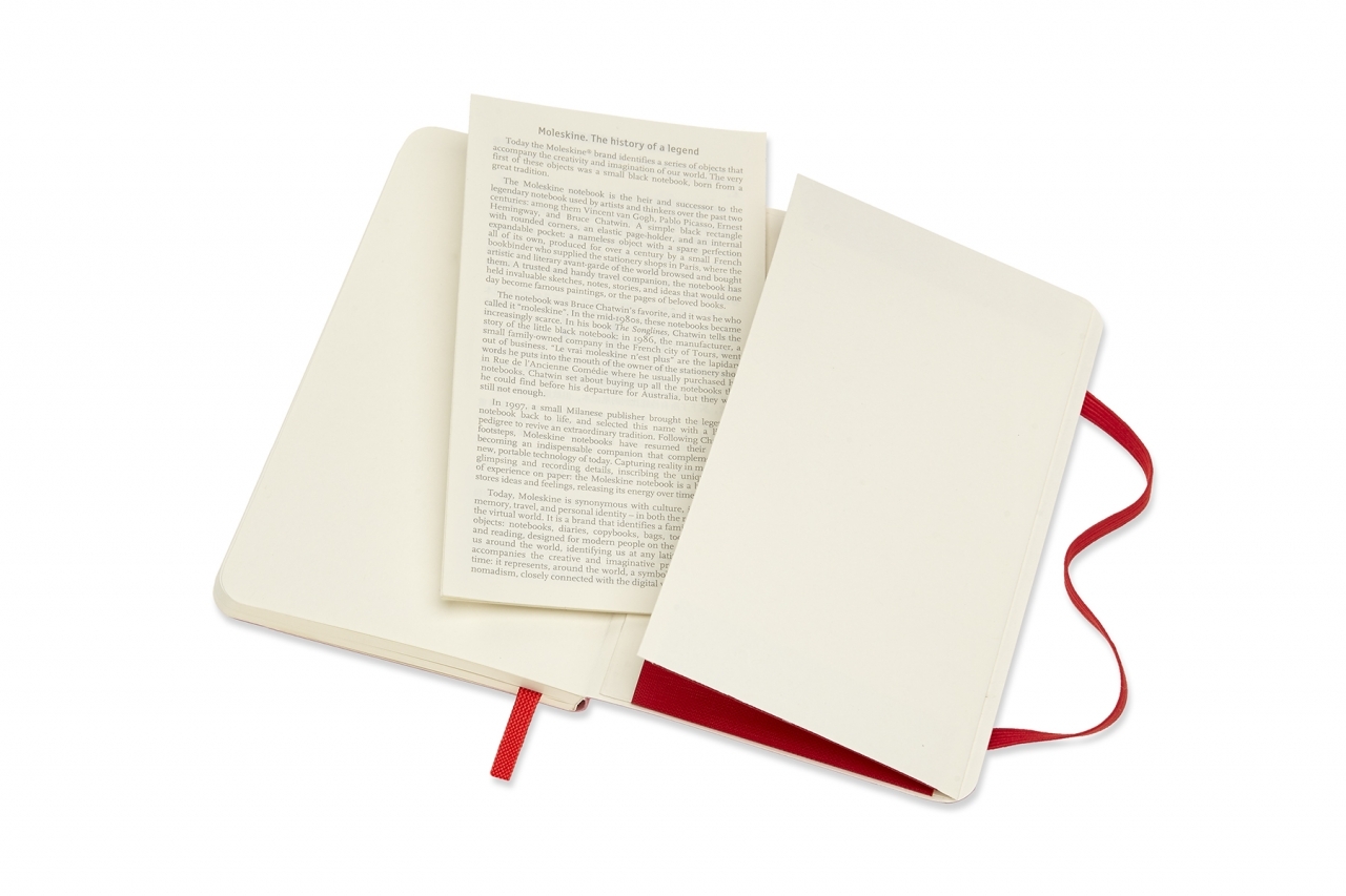 Atversta knygutė su kreminiais lapais, tarp jų įdėtas Moleskine The history of a legend tekstas, apačioje raudonas viršelis