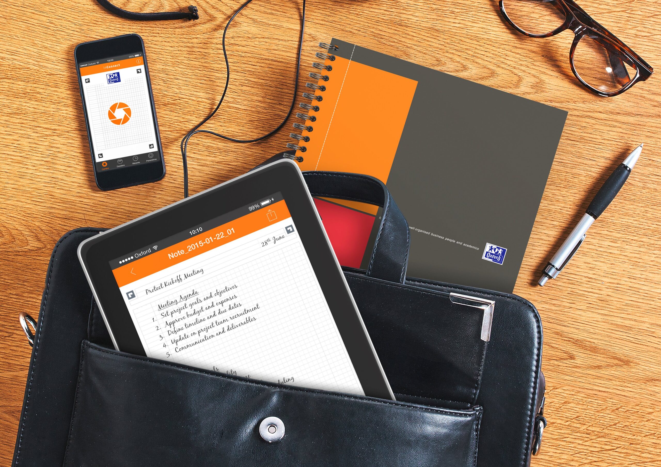 Oranžinis Oxford Activebook sąsiuvinis šalia planšetės krepšyje, telefono su Scribzee ženklu, rašiklio, akinių ir ausinių
